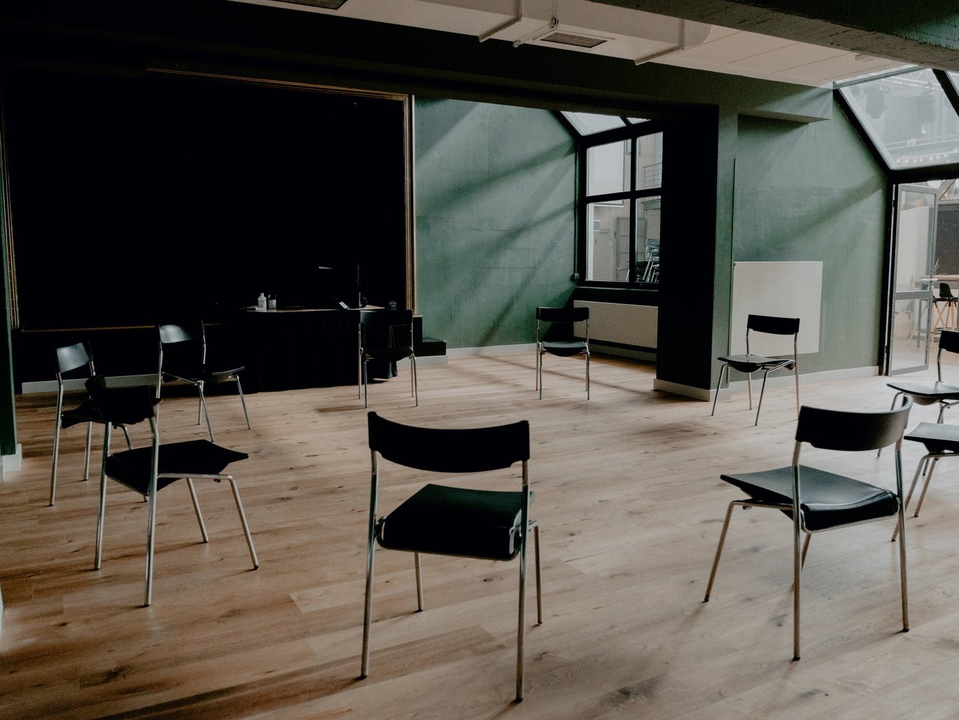 Zu sehen ist ein kleiner Theatersaal mit Tannengrünen Wänden und hellem Holzfußboden. Im Hintergrund ist eine dunkle Bühne zu erkennen. Den Vordergrund dominiert ein Stuhlkreis.