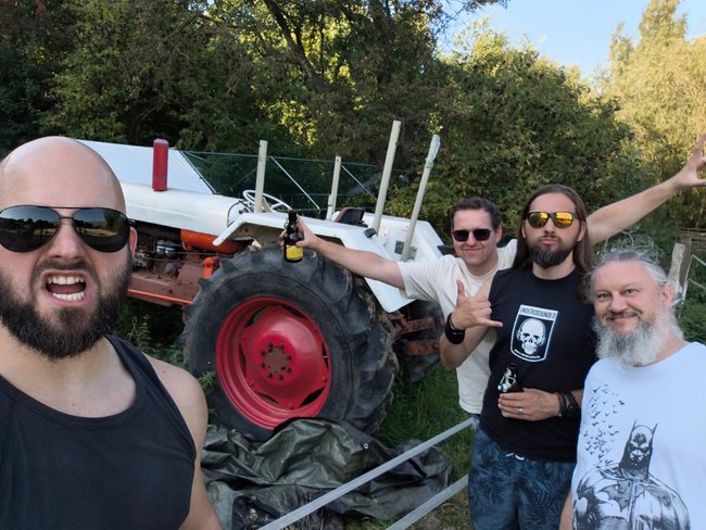 Auf dem Bild sieht man die Band Blackrout. Sie stehen vor und neben einem Trecker. Im Hintergrund sieht man eine Hecke und blauen Himmel. Auf einen Teil der Hecke scheint die Sonne.