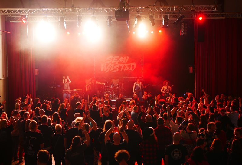 Zu sehen ist der Saal der WERK°STADT während eines Metal Konzertes von Semi Wasted. Das Bild ist von hinten aus dem Publikum aufgenommen worden. Die Bühne ist in roten Farben ausgeleuchtet. Die 5 Bandmitglieder performen vor einem großen Publikum. 